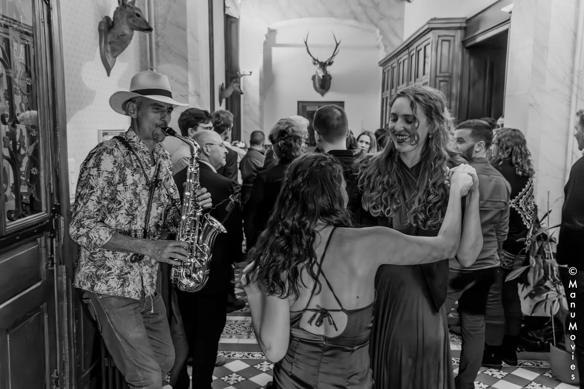 Saxophoniste de cocktail de mariage jouant dans le hall d'un château pendant que les invités dansent (photo noir et blanc)